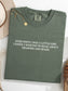 A moss green folded t-shirt with white printed text reading 'EVER SINCE I WAS A LITTLE GIRL I KNEW I WANTED TO READ ABOUT DRAGONS AND MAGIC', styled on a white surface with a gold necklace and a beige hat partially visible.