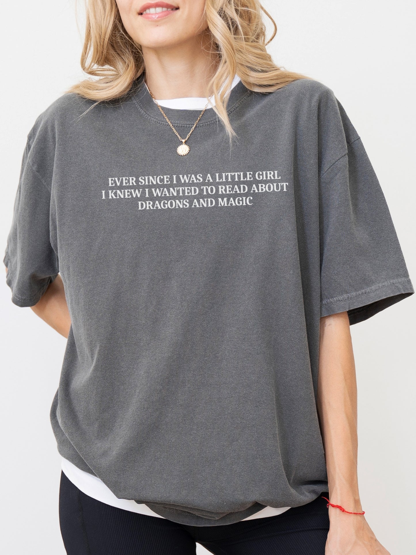 A woman wearing an oversized pepper gray t-shirt with white text that reads 'EVER SINCE I WAS A LITTLE GIRL, I KNEW I WANTED TO READ ABOUT DRAGONS AND MAGIC'.