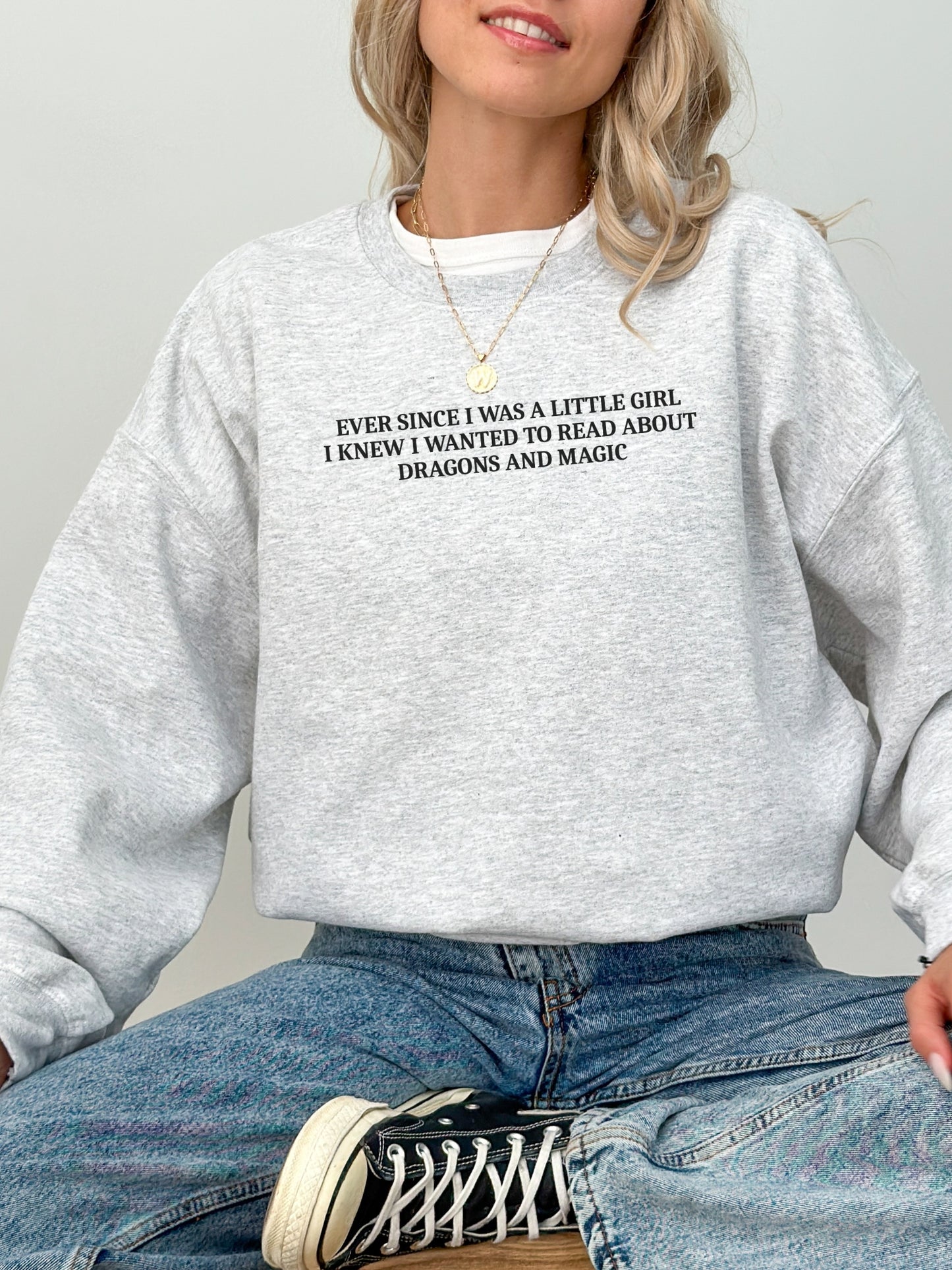 A woman wearing an ash heather gray crewneck sweatshirt with black text that reads 'EVER SINCE I WAS A LITTLE GIRL, I KNEW I WANTED TO READ ABOUT DRAGONS AND MAGIC'. She is also wearing blue jeans and a black and white canvas sneaker.
