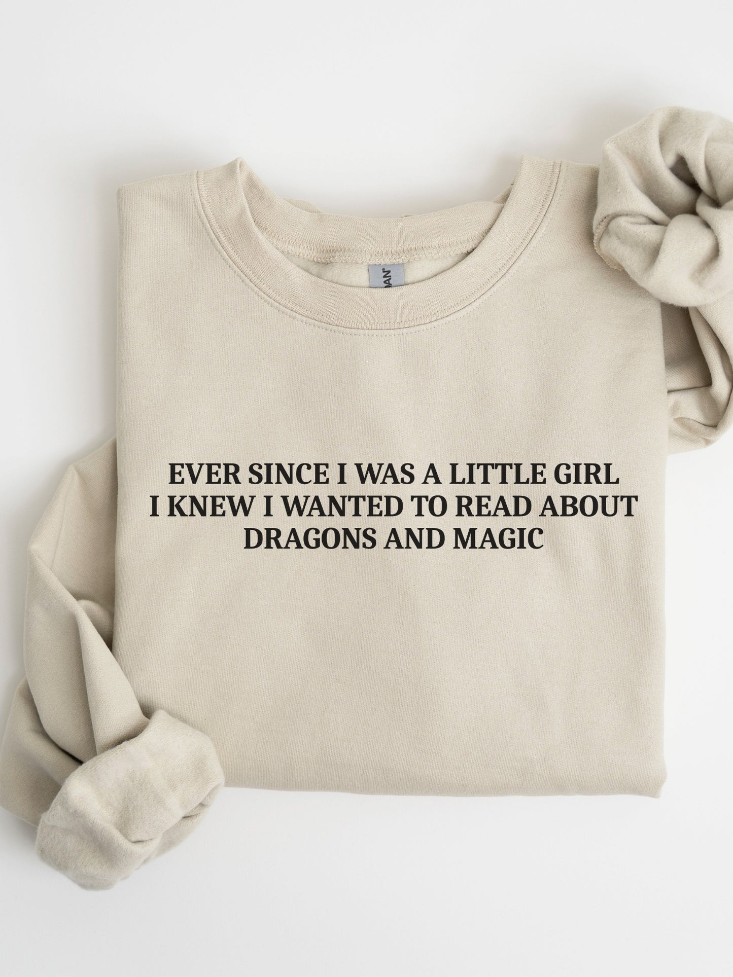 A folded sand crewneck sweatshirt with black text reading 'EVER SINCE I WAS A LITTLE GIRL I KNEW I WANTED TO READ ABOUT DRAGONS AND MAGIC', styled with a matching fabric scrunchie on a white background.