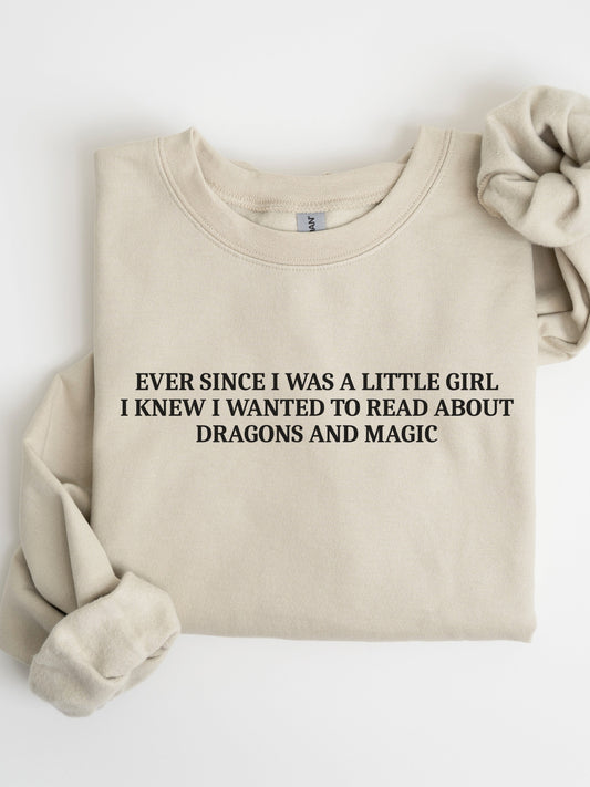 A folded sand crewneck sweatshirt with black text reading 'EVER SINCE I WAS A LITTLE GIRL I KNEW I WANTED TO READ ABOUT DRAGONS AND MAGIC', styled with a matching fabric scrunchie on a white background.