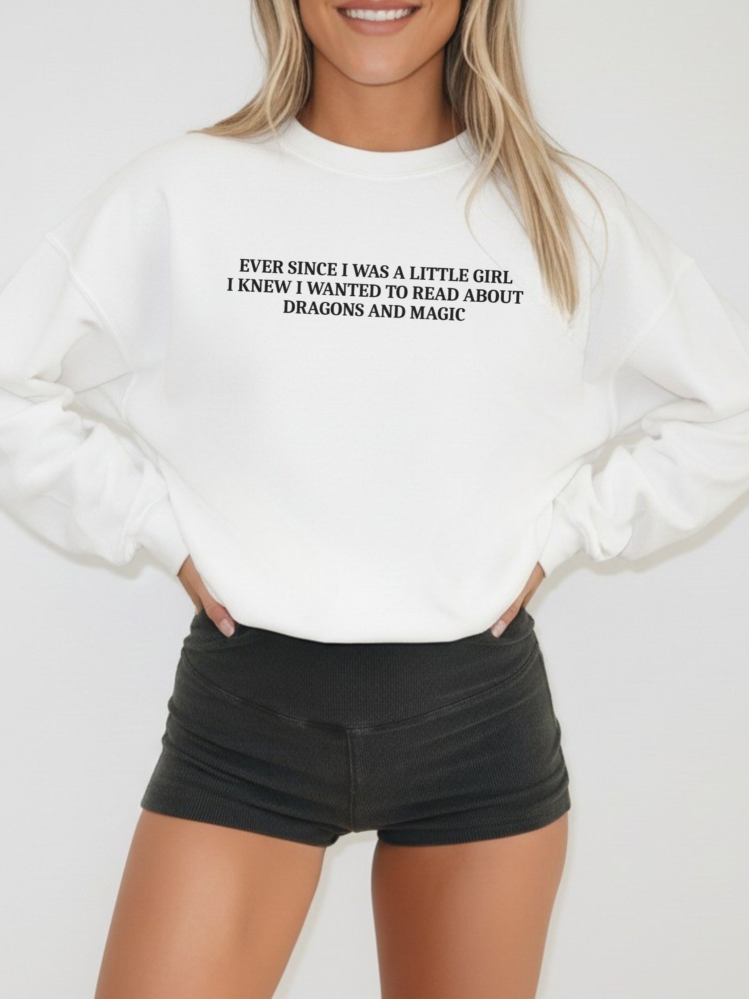 A woman wearing a white crewneck sweatshirt with black text that reads 'EVER SINCE I WAS A LITTLE GIRL, I KNEW I WANTED TO READ ABOUT DRAGONS AND MAGIC', paired with black shorts, against a plain light gray background.