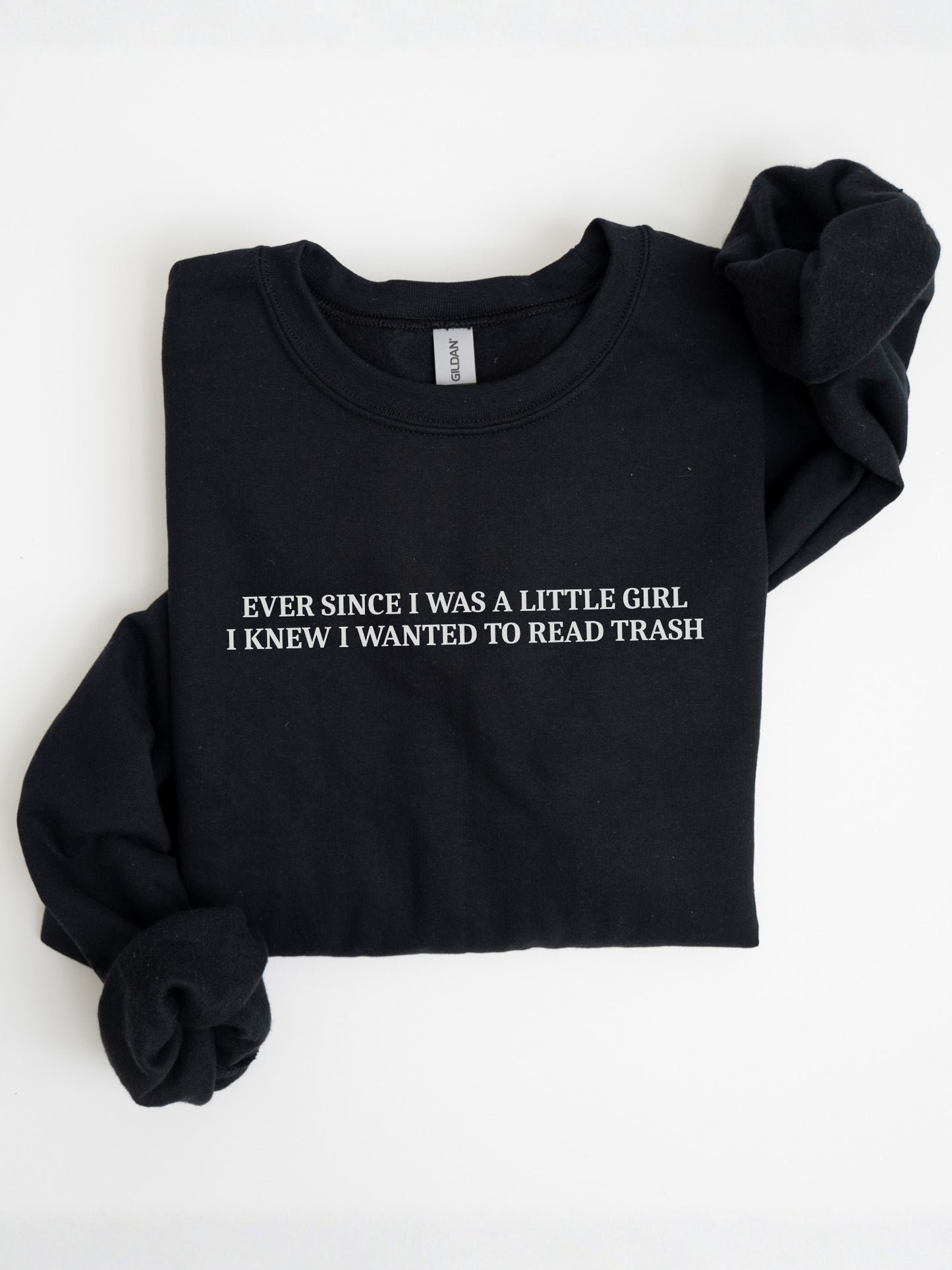 A navy blue folded crewneck sweatshirt with white text reading 'EVER SINCE I WAS A LITTLE GIRL. I KNEW I WANTED TO READ TRASH' on the chest, displayed on a clean white background.
