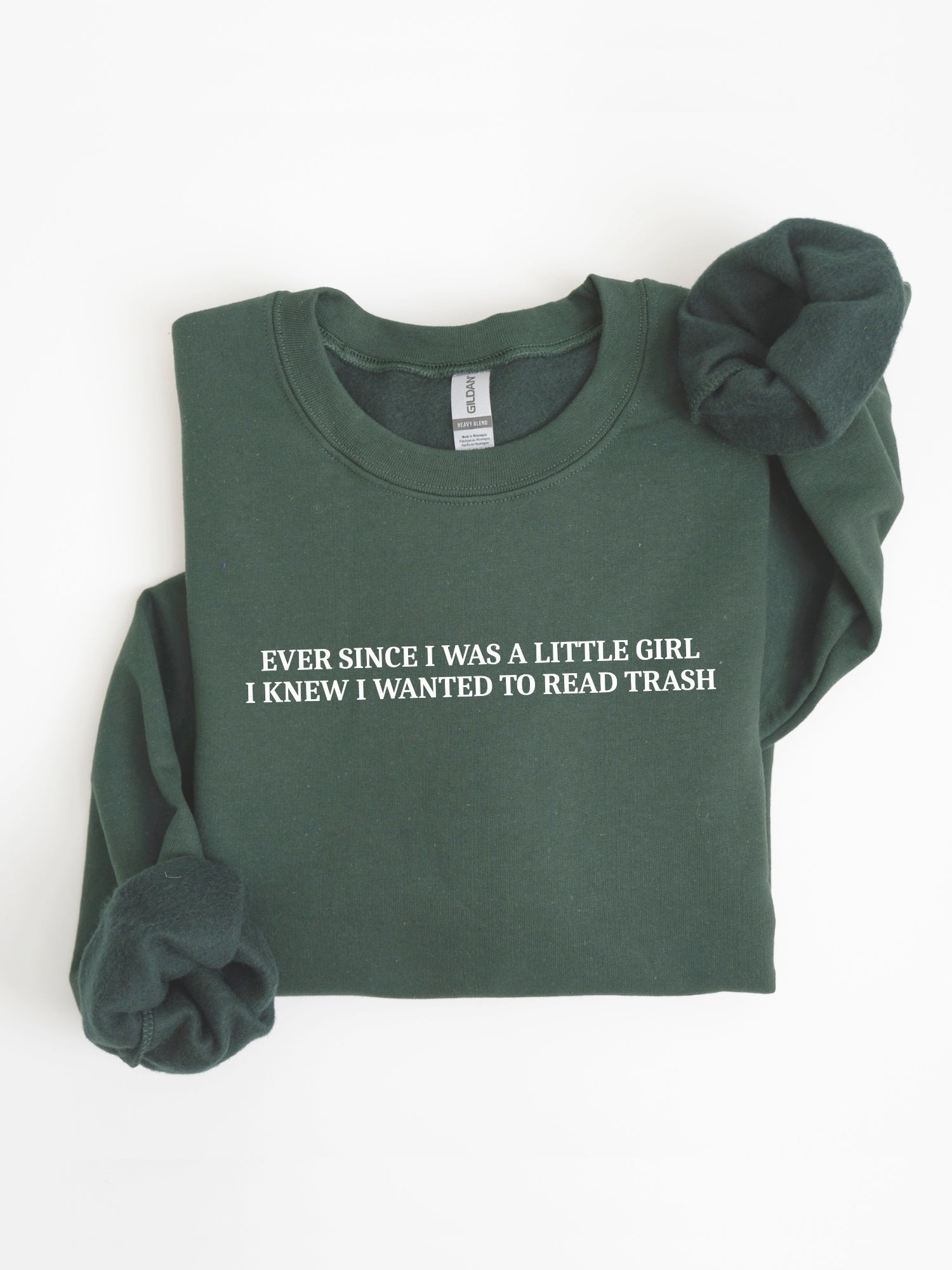 A dark forest green crewneck sweatshirt with white text reading 'EVER SINCE I WAS A LITTLE GIRL, I KNEW I WANTED TO READ TRASH' on the chest, shown folded on a white background with rolled cuffs.