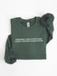 A dark forest green crewneck sweatshirt with white text reading 'EVER SINCE I WAS A LITTLE GIRL, I KNEW I WANTED TO READ TRASH' on the chest, shown folded on a white background with rolled cuffs.