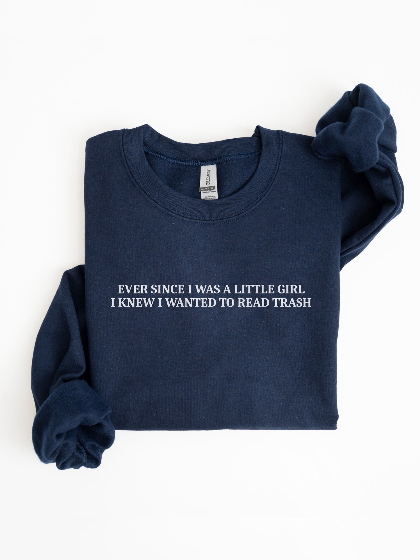 A navy blue folded crewneck sweatshirt with white text reading 'EVER SINCE I WAS A LITTLE GIRL. I KNEW I WANTED TO READ TRASH' on the chest, displayed on a clean white background.