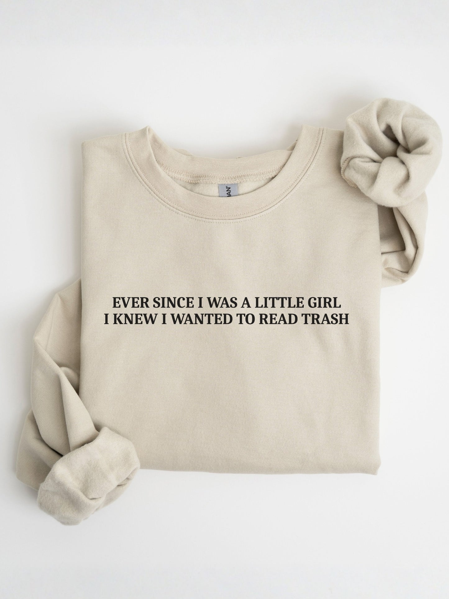 A folded sand crewneck sweatshirt with the text 'EVER SINCE I WAS A LITTLE GIRL I KNEW I WANTED TO READ TRASH' printed in black on the chest. A matching beige fabric scrunchie is placed on the upper right shoulder, all set against a clean white background.