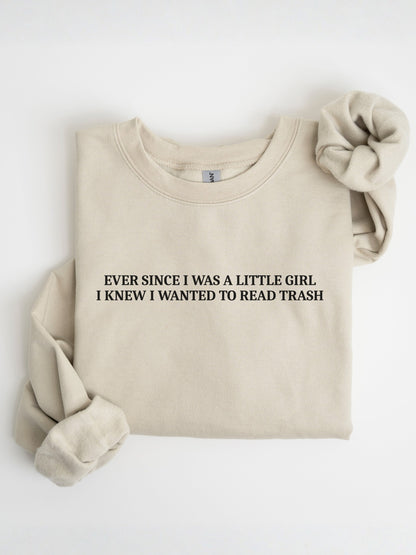 A folded sand crewneck sweatshirt with the text 'EVER SINCE I WAS A LITTLE GIRL I KNEW I WANTED TO READ TRASH' printed in black on the chest. A matching beige fabric scrunchie is placed on the upper right shoulder, all set against a clean white background.