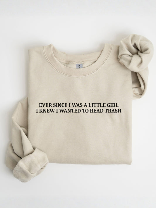 A folded sand crewneck sweatshirt with the text 'EVER SINCE I WAS A LITTLE GIRL I KNEW I WANTED TO READ TRASH' printed in black on the chest. A matching beige fabric scrunchie is placed on the upper right shoulder, all set against a clean white background.