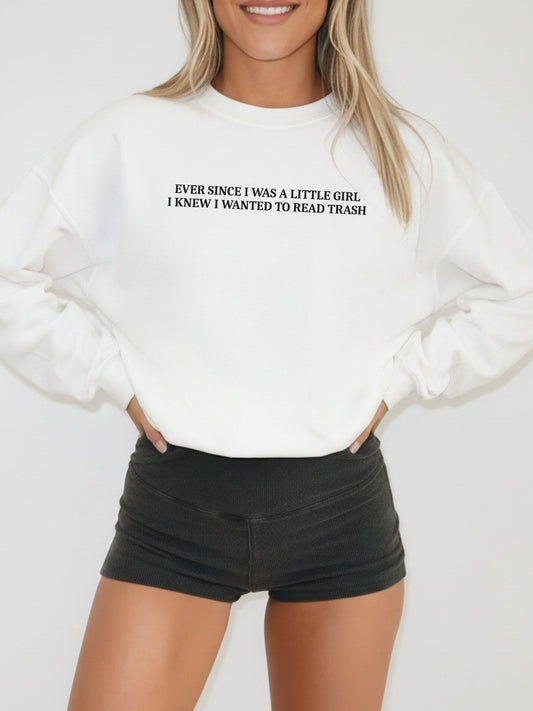 A woman wearing a white crewneck sweatshirt with black text that reads 'EVER SINCE I WAS A LITTLE GIRL, I KNEW I WANTED TO READ TRASH!' paired with black denim shorts, against a plain light gray background.