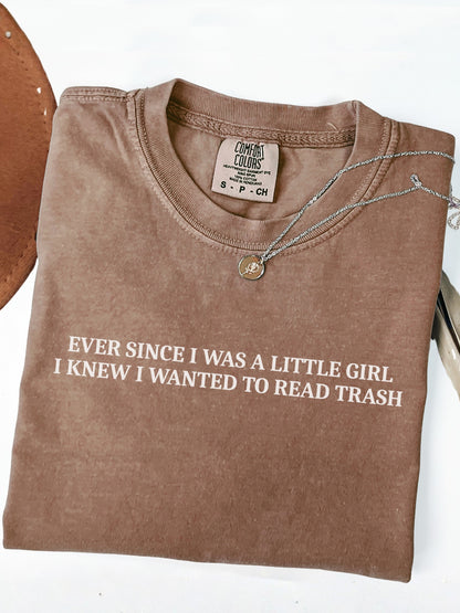 A folded espresso brown t-shirt with white text reading 'EVER SINCE I WAS A LITTLE GIRL I KNEW I WANTED TO READ TRASH' on a white background, styled with a necklace and a leather object edge.