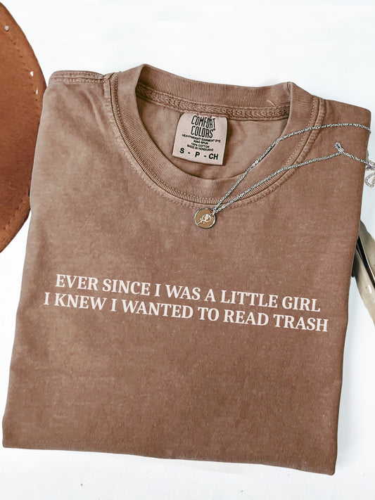 A folded espresso brown t-shirt with white text reading 'EVER SINCE I WAS A LITTLE GIRL I KNEW I WANTED TO READ TRASH' on a white background, styled with a necklace and a leather object edge.