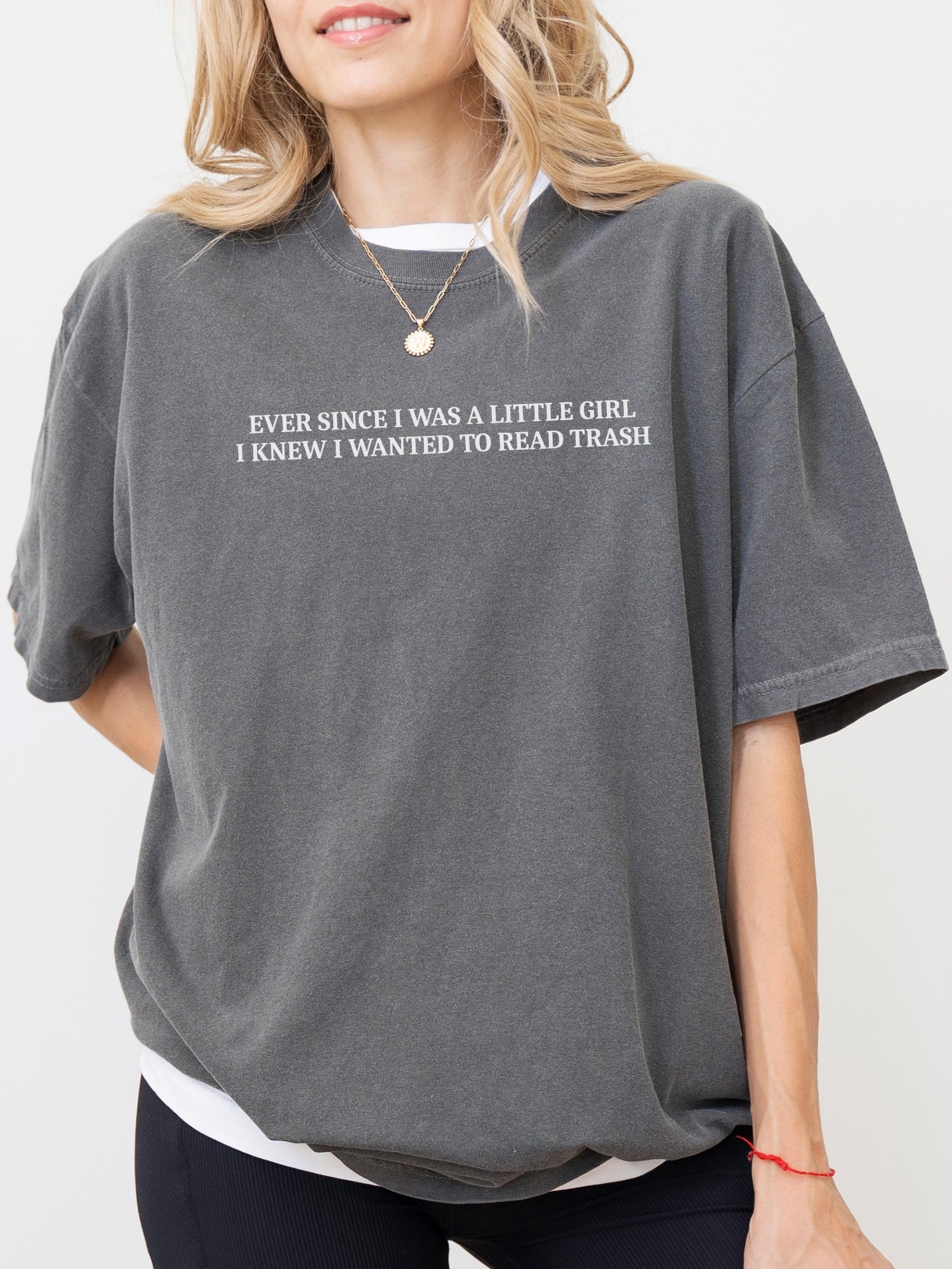 A woman wearing an oversized pepper graphic t-shirt with white text that reads 'EVER SINCE I WAS A LITTLE GIRL, I KNEW I WANTED TO READ TRASH'.