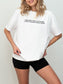 A woman wearing an oversized white t-shirt with a black graphic print that reads "Ever Since I Was a Little Girl I Knew I Wanted to Read Trash". She is also wearing black shorts and a delicate gold necklace, set against a plain light gray background.