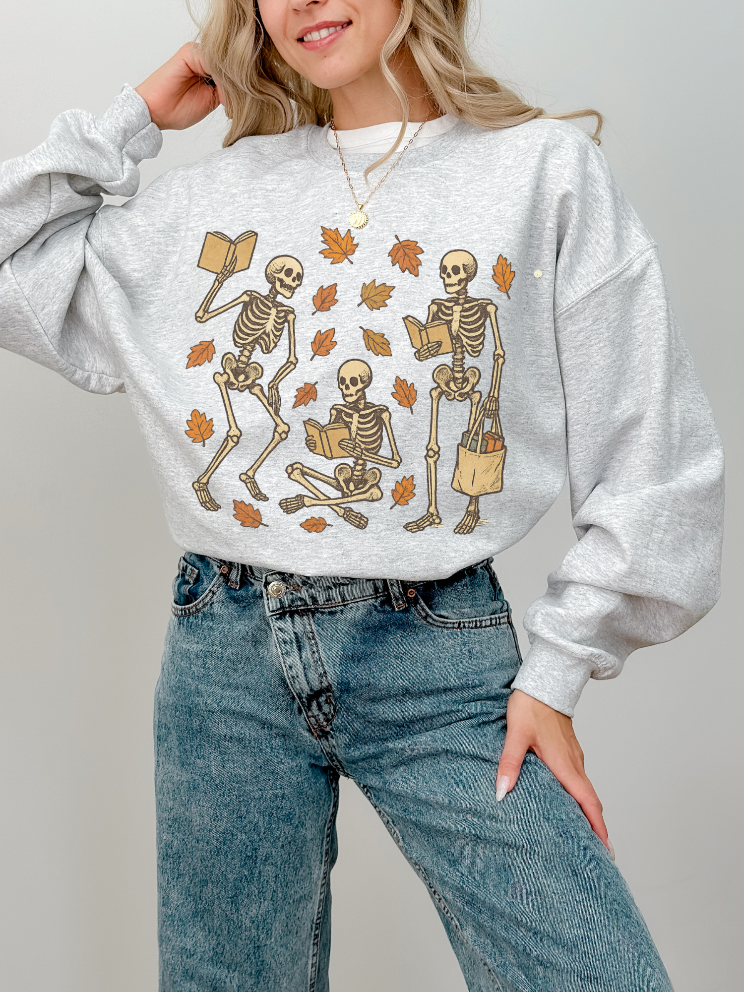Light gray sweatshirt with cozy autumn bookish skeletons design, worn by a woman standing with hand on hip and smiling