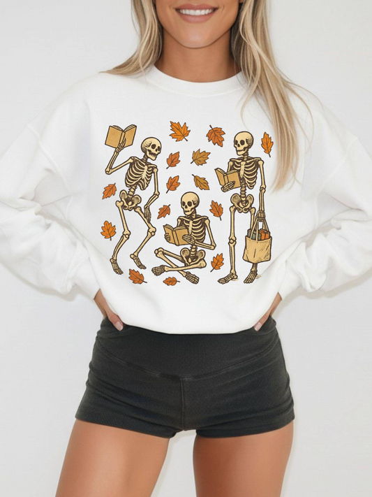 Woman in white sweatshirt featuring three skeletons reading books and holding a tote, surrounded by orange and brown leaves, styled with black shorts