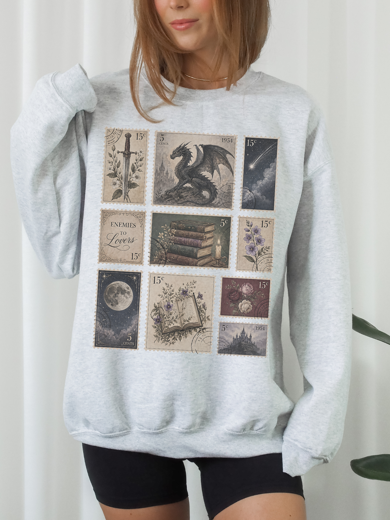 Woman wearing ash gray sweatshirt with vintage postage stamp collage featuring dragon sword books moon flowers castle and enemies to lovers fantasy book design