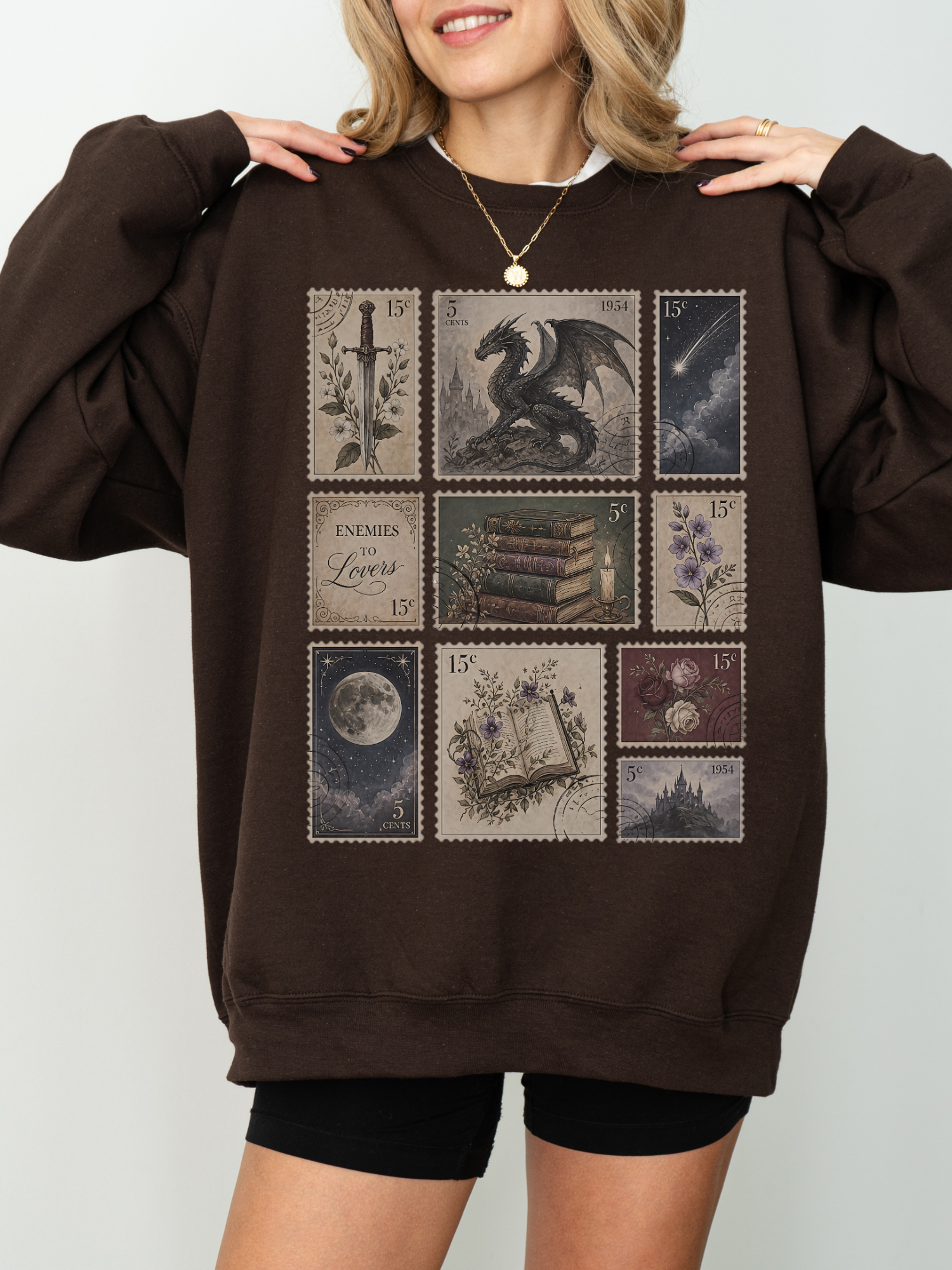 Woman wearing brown oversized sweatshirt with vintage fantasy stamp collage featuring dragon sword books moon floral art and enemies to lovers inspired design