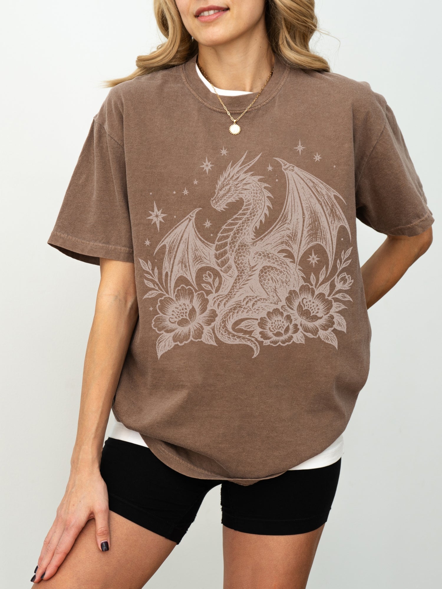 A woman wearing a espresso short-sleeve t-shirt with a large white dragon and floral graphic on the front, paired with black shorts, against a light gray studio background.