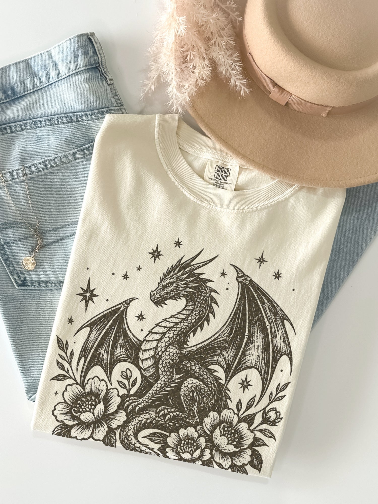 A folded ivory-colored t-shirt featuring a detailed black dragon illustration with flowers and stars, styled in a flat lay with light-wash denim jeans, a beige hat, and dried feathers on a white background.
