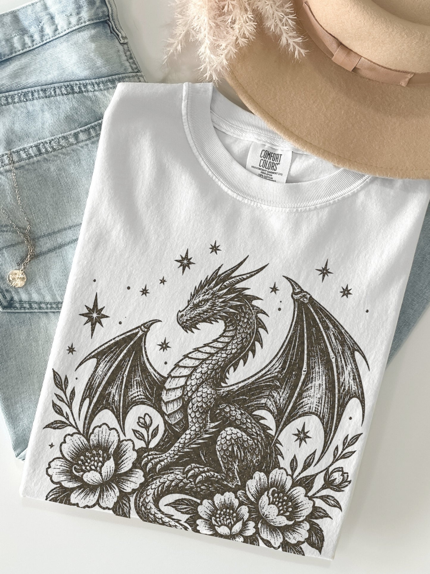 A folded white T-shirt with a detailed black dragon illustration, surrounded by flowers and stars, styled with light-wash denim shorts and a beige hat on a light background.