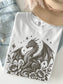 A folded white T-shirt with a detailed black dragon illustration, surrounded by flowers and stars, styled with light-wash denim shorts and a beige hat on a light background.