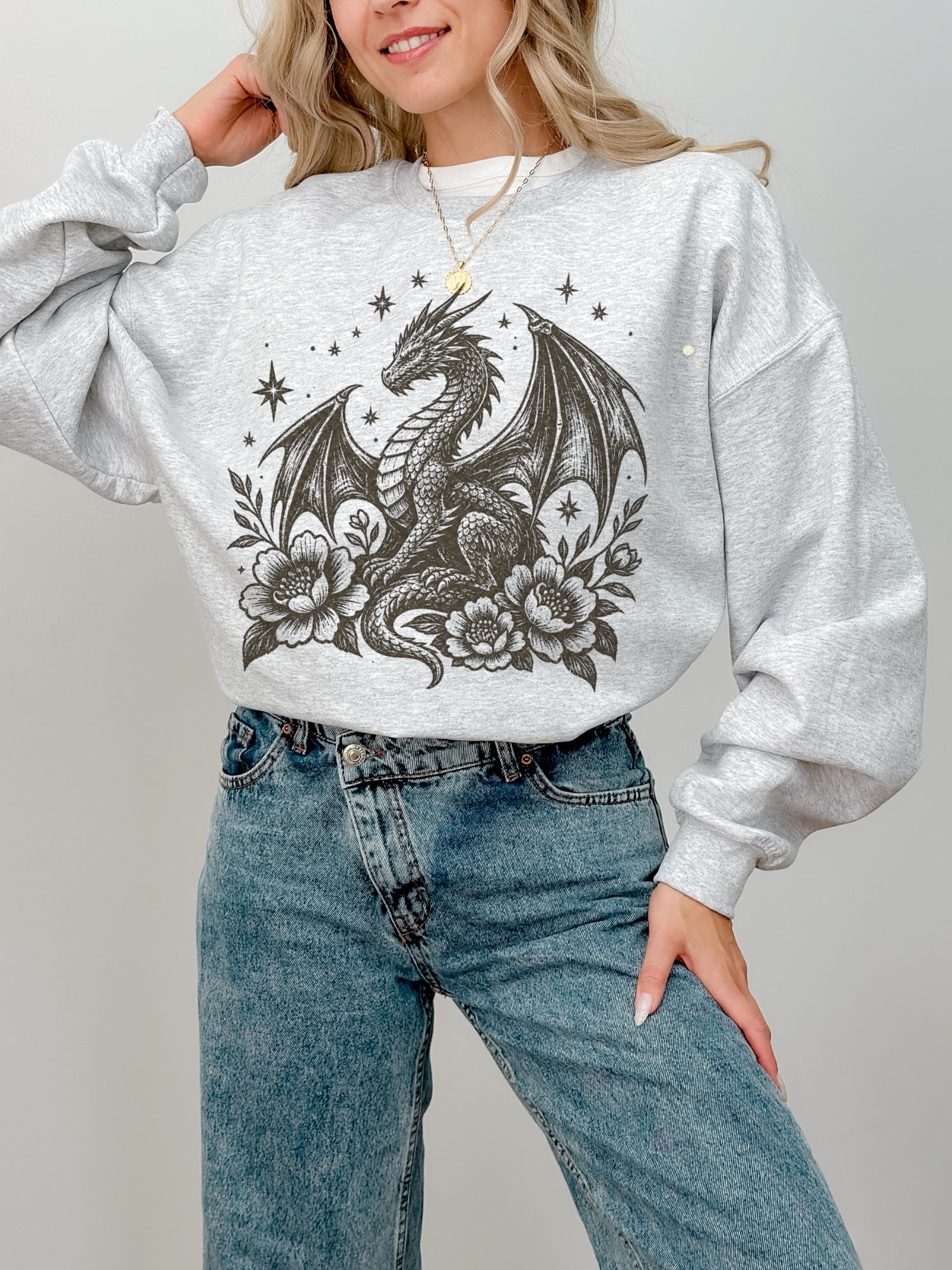 A woman wearing an ash gray heather crewneck sweatshirt with a black graphic of a dragon surrounded by flowers and stars, paired with blue jeans.