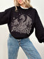 A woman wearing a black crewneck sweatshirt with a detailed gray dragon and floral graphic on the front, paired with light wash blue jeans.