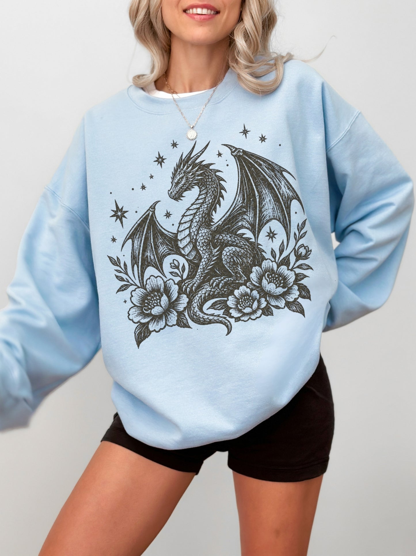 A woman wearing a light blue crewneck sweatshirt with a black graphic of a dragon surrounded by flowers and stars, paired with black shorts.