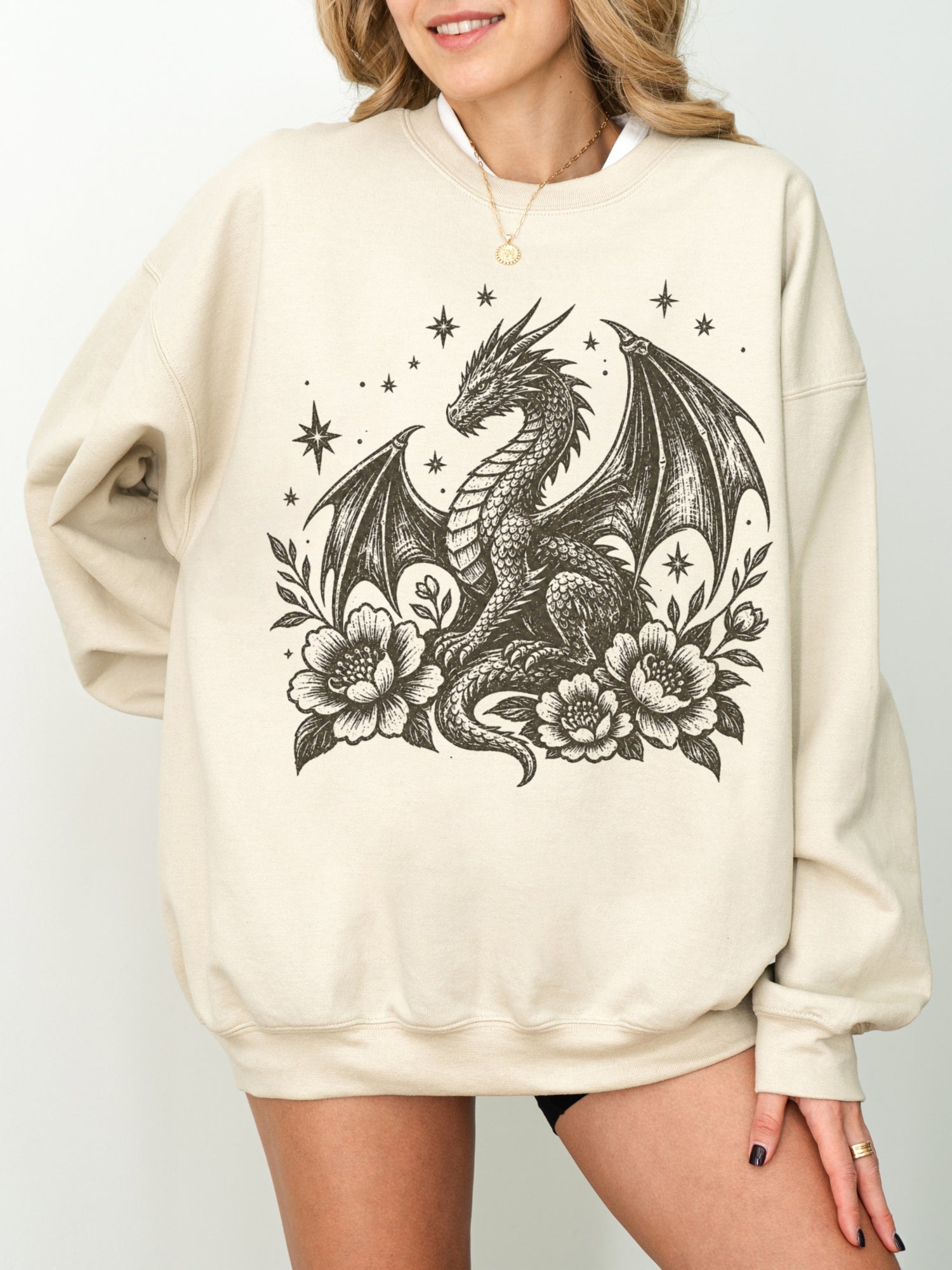 A woman wearing an oversized sand-colored sweatshirt with a detailed black graphic of a dragon surrounded by flowers and stars.