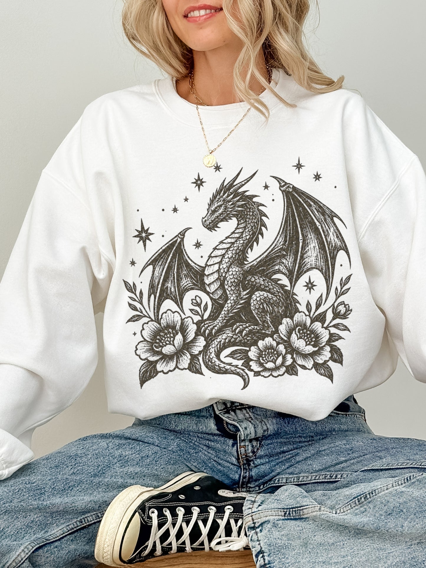 A woman wearing a white crewneck sweatshirt with a black dragon and floral graphic, paired with blue jeans and a black Converse sneaker.