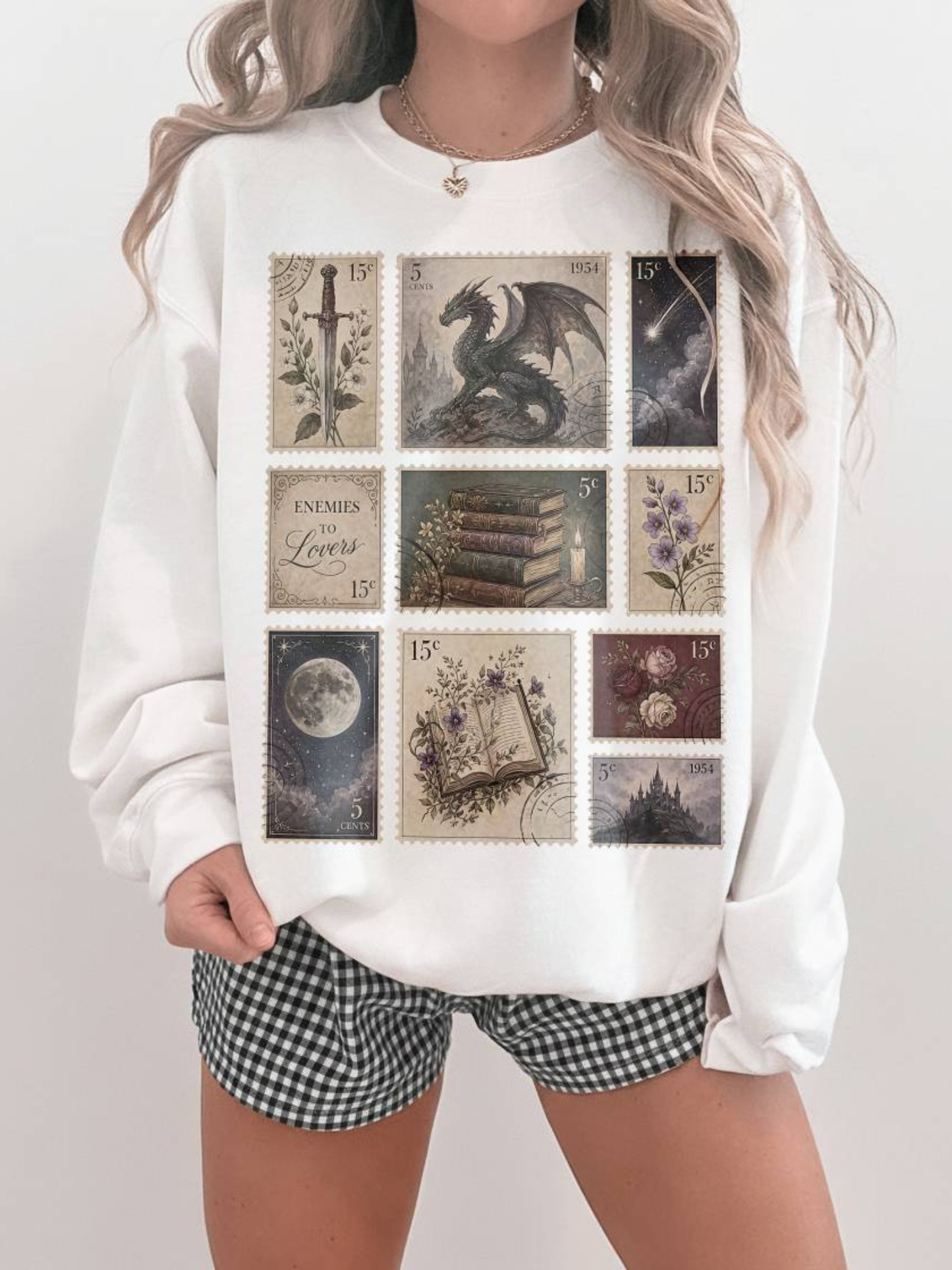 Woman wearing white oversized sweatshirt featuring vintage fantasy postage stamp illustrations with dragon moon books flowers castle and enemies to lovers aesthetic paired with gingham shorts