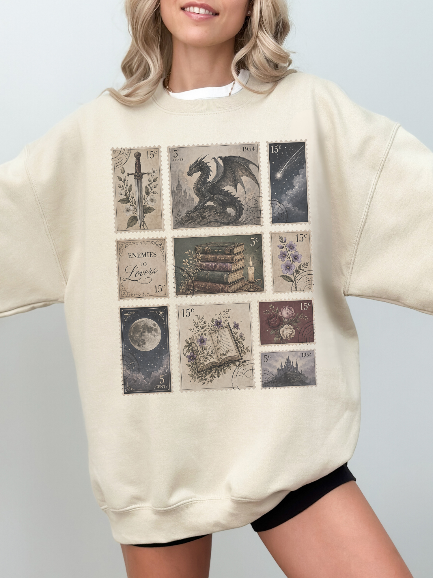 Woman wearing sand oversized sweatshirt with fantasy themed postage stamp artwork including dragon antique books moon floral illustrations and castle aesthetic