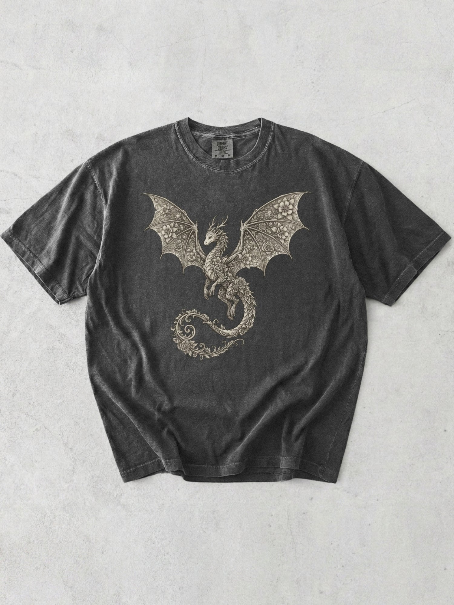A pepper t-shirt featuring a detailed graphic of a dragon with spread wings in a metallic gold/bronze color, laid flat on a light gray textured background.