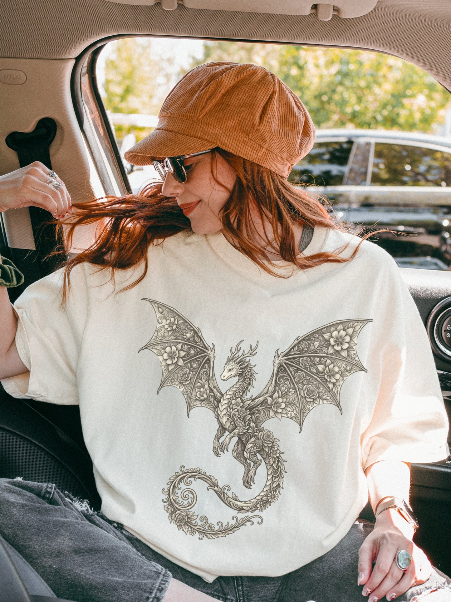 A woman sitting inside a car wearing an ivory-colored t-shirt with a detailed gray dragon graphic on the front, paired with a brown corduroy cap and sunglasses.