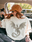 A woman sitting inside a car wearing an ivory-colored t-shirt with a detailed gray dragon graphic on the front, paired with a brown corduroy cap and sunglasses.