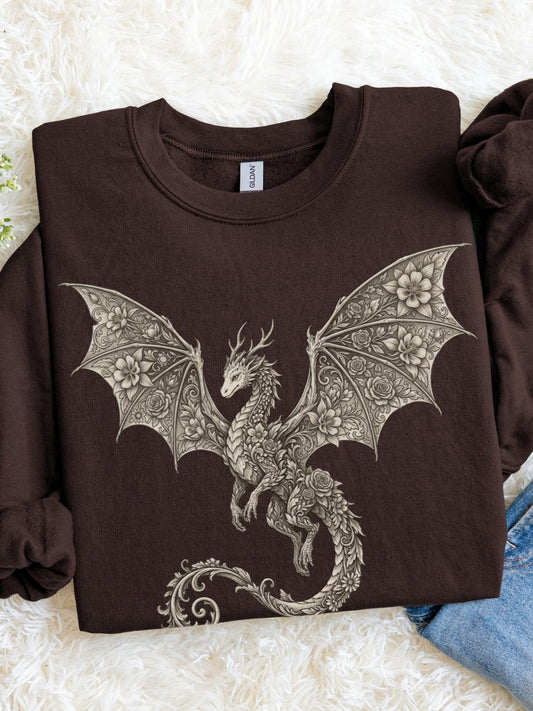 A dark brown crewneck sweatshirt featuring a large, detailed gray dragon graphic with intricate wing patterns, laid flat on a white faux fur surface with a blue denim garment partially visible at the bottom right.