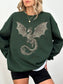 A woman wearing an oversized forest green sweatshirt with a large silver dragon graphic on the front, paired with black shorts and a gold necklace, against a plain white background.