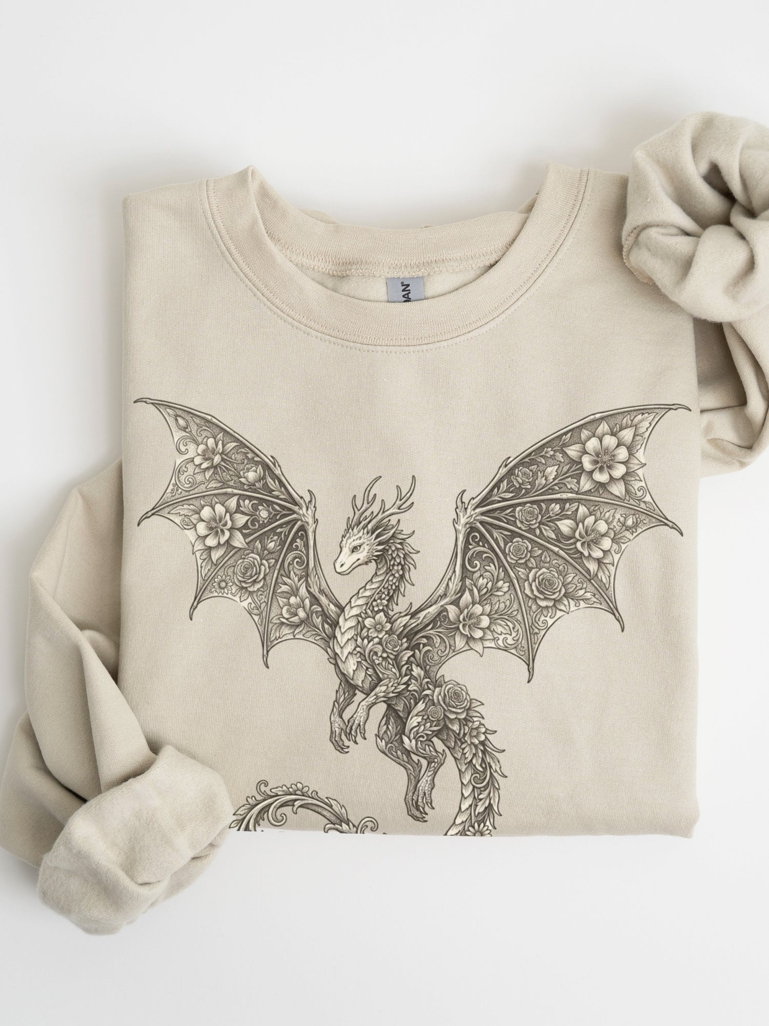 A folded sand crewneck sweatshirt featuring a detailed black line art graphic of a dragon with intricate wing patterns, styled with a matching fabric scrunchie on a white background.