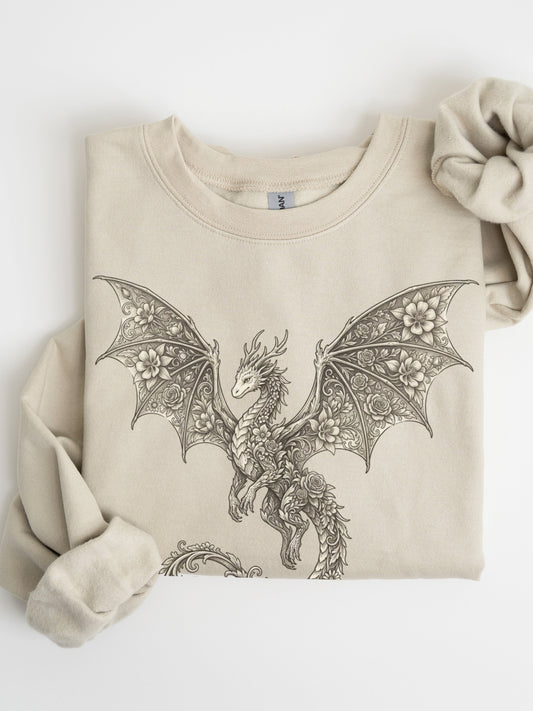 A folded sand crewneck sweatshirt featuring a detailed black line art graphic of a dragon with intricate wing patterns, styled with a matching fabric scrunchie on a white background.