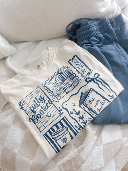 Ivory fully booked book lover t shirt with blue vintage style reading graphics folded on bed next to blue sweatpants cozy aesthetic flatlay