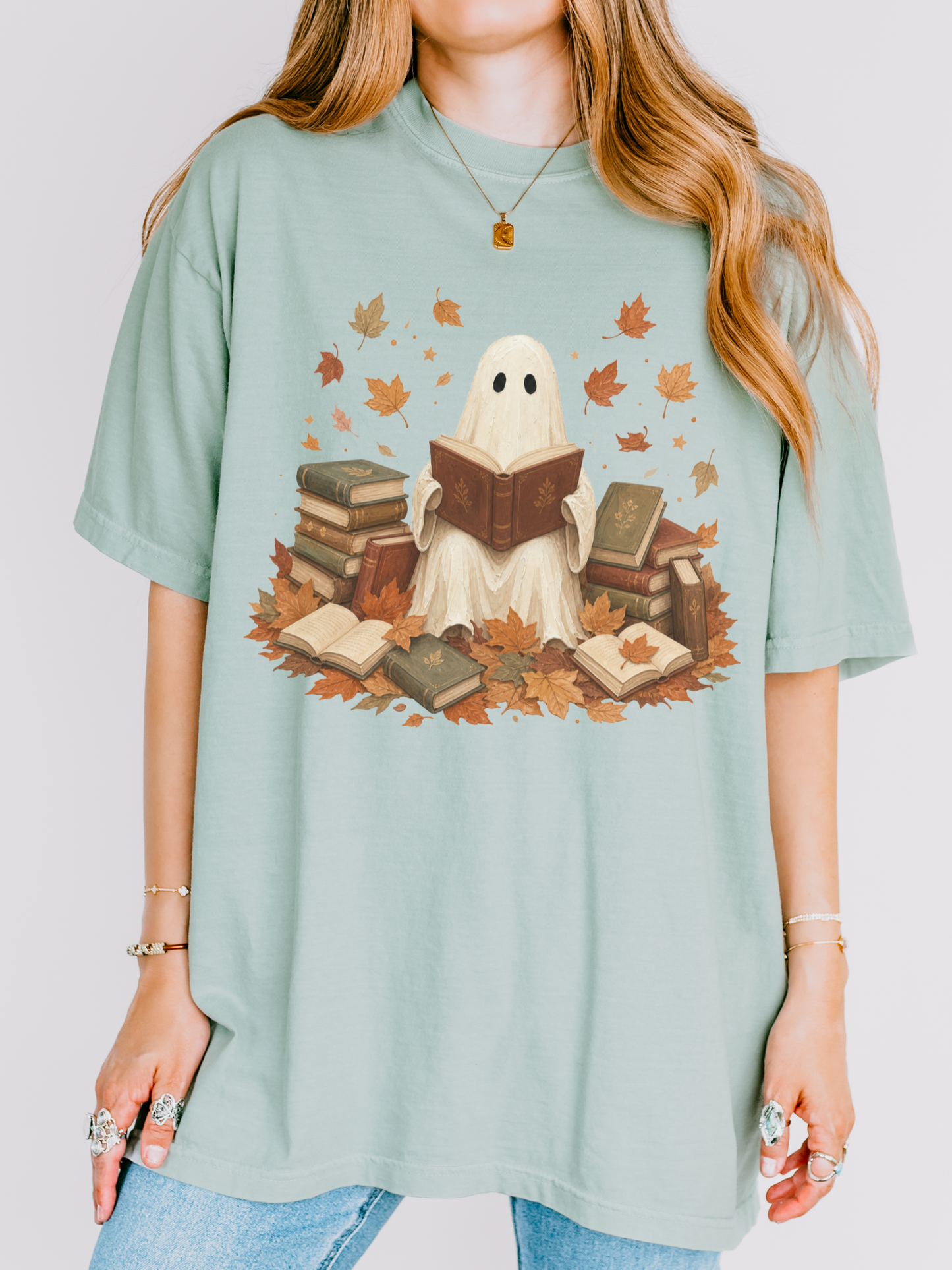 A woman wearing an oversized bay green t-shirt with a graphic of a ghost reading books surrounded by autumn leaves, paired with blue jeans.