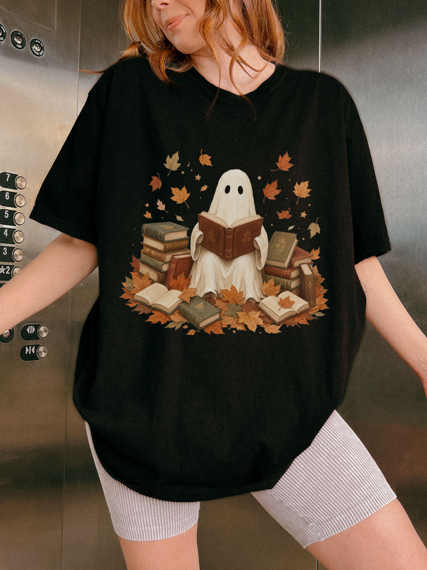 A woman wearing an oversized black graphic t-shirt with a cute ghost reading books design, paired with white ribbed shorts, standing in front of a metallic elevator wall.