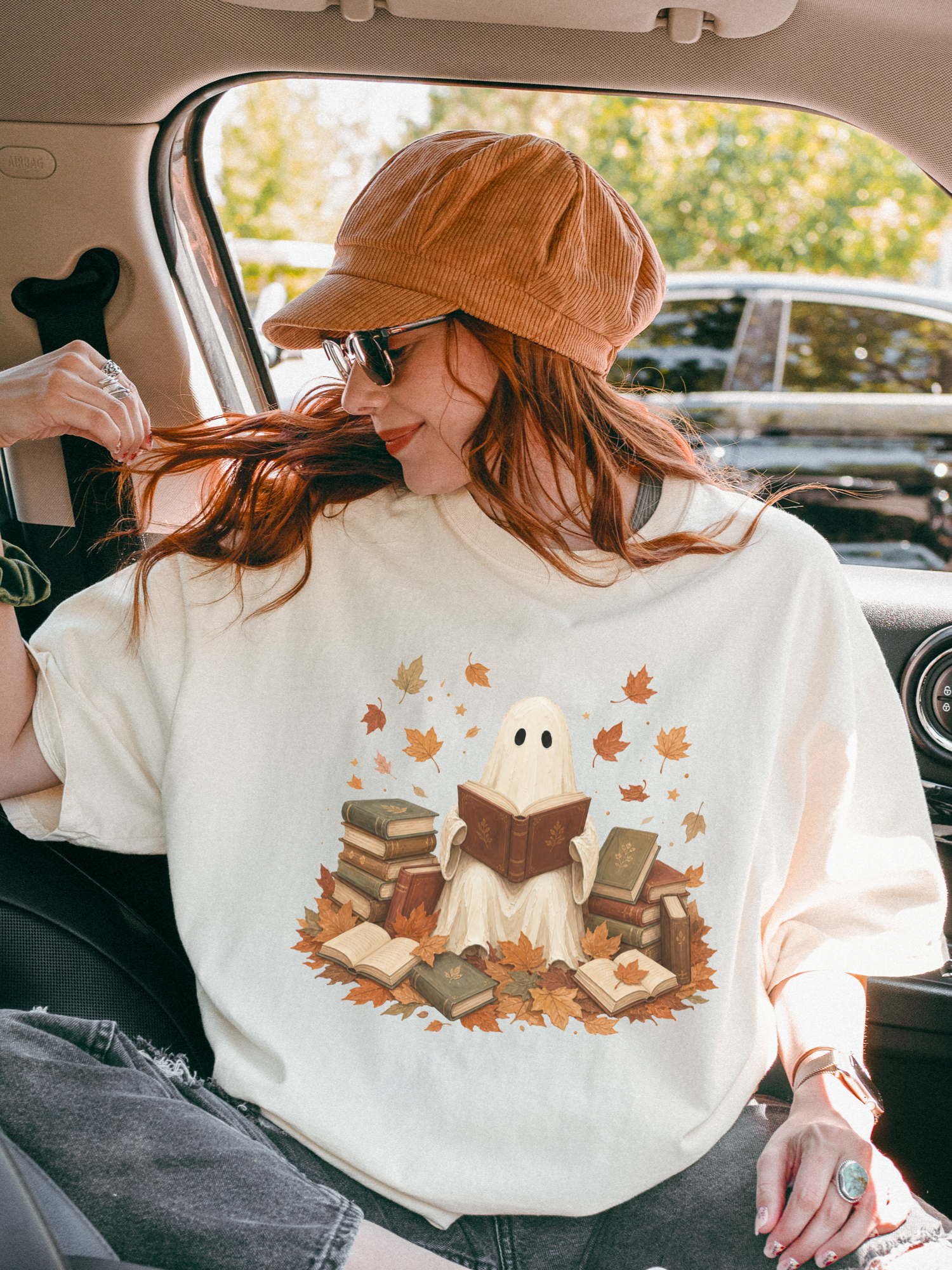 A woman sitting inside a car wearing an ivory t-shirt with a ghost reading books design, styled with a brown corduroy cap and sunglasses.