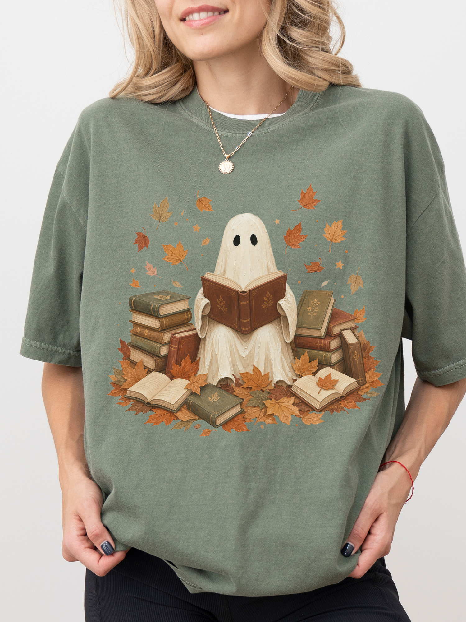 A woman wearing an oversized moss green t-shirt with a graphic of a ghost reading a book surrounded by autumn leaves and stacks of old books.