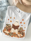 A white graphic T-shirt laid flat on a light surface, styled with blue denim jeans, a tan hat, and a necklace. The shirt features a printed illustration of a ghost-like figure reading a book surrounded by autumn leaves and stacks of books.