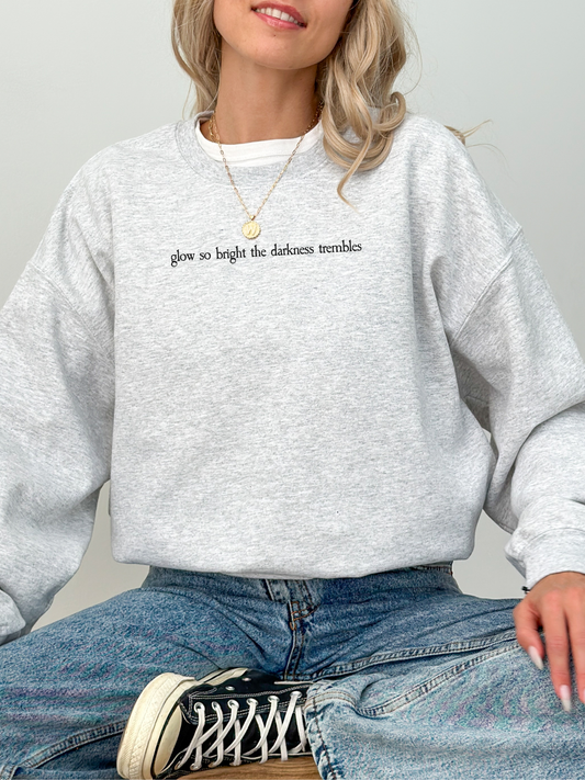 Woman wearing a light heather ash Gildan sweatshirt that says "glow so bright the darkness trembles" in black serif text across the chest. She is sitting cross-legged in denim jeans and sneakers, smiling confidently.