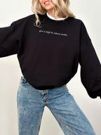 Woman wearing a black Gildan crewneck with white serif text reading "glow so bright the darkness trembles." She is mid-laugh with arms open in a playful pose, styled with classic jeans and layered necklaces.

