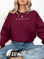 Woman wearing a maroon Gildan crewneck featuring the phrase “glow so bright the darkness trembles” in white serif font. She is seated in a relaxed pose with light denim and black Converse sneakers.

