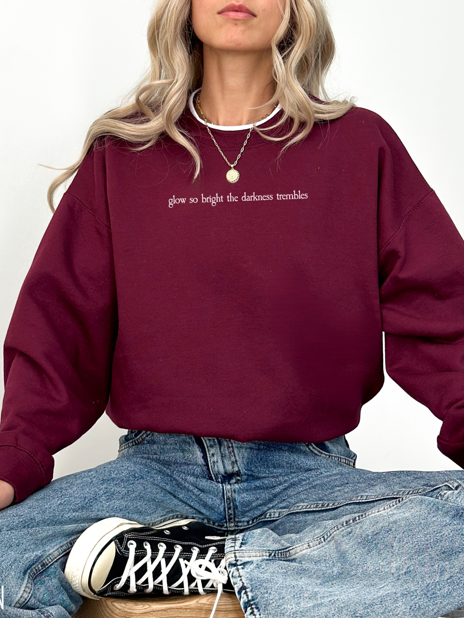 Woman wearing a maroon Gildan crewneck featuring the phrase “glow so bright the darkness trembles” in white serif font. She is seated in a relaxed pose with light denim and black Converse sneakers.


